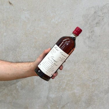 A hand holding a bottle of Michel Couvreur Peated Overaged Whisky with a label that includes the brand name and the product name, against a grey concrete background.