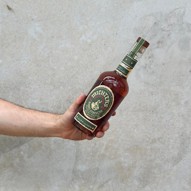 A hand holding a bottle of Michter's Limited Edition Barrel Strength Rye Whisky with a concrete surface in the background.