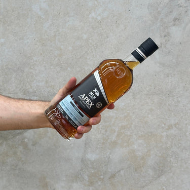 A hand holding a bottle of Milk & Honey APEX Dead Sea Whisky with a label and clear liquid visible inside the bottle.