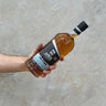 A hand holding a bottle of Milk & Honey APEX Dead Sea Whisky with a label and clear liquid visible inside the bottle.