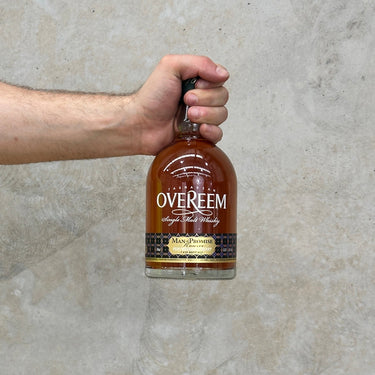 A hand holding a bottle of Overeem Man Of Promise 2022 Whisky with a label and clear liquid visible inside the bottle.