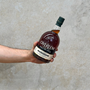 A hand holding a bottle of Overeem Sherry Cask - Cask Strength Whisky with a label and cap, against a grey background.