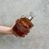 A hand holding a square-shaped decanter of Overeem XO Brandy with a wooden top and a label on the front.
