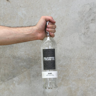 A hand holding a bottle of Pasote Blanco tequila with a concrete background.