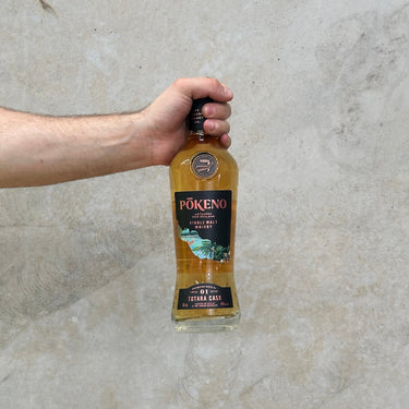 A hand holding a bottle of Pokeno Exploration Series No. 1 Totara Barrel Whisky against a grey concrete background.