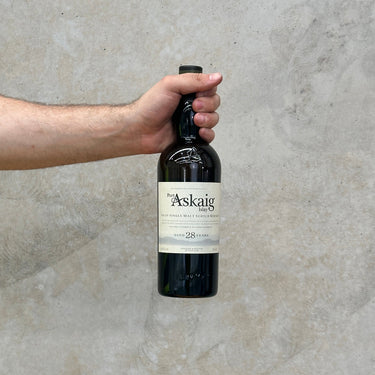 A hand holding a bottle of Port Askaig 28 Year Old Whisky against a grey concrete background.