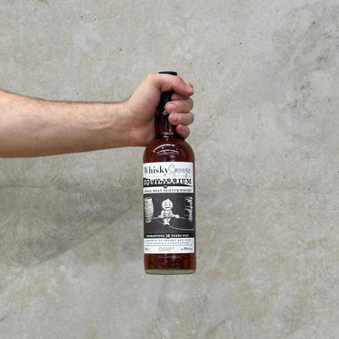A hand holding a bottle of Edradour Whisky Sponge Equilibrium II Edradour & Ballechin 16 Year Old whisky against a grey background.