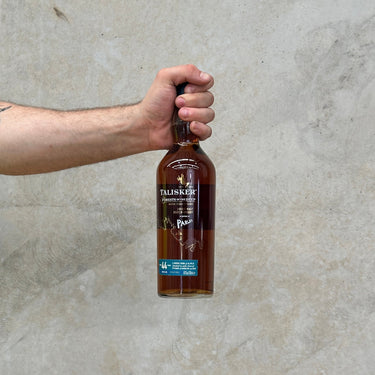 A hand holding a bottle of Talisker Forests Of The Deep 44 Year Old Whisky against a grey concrete background.