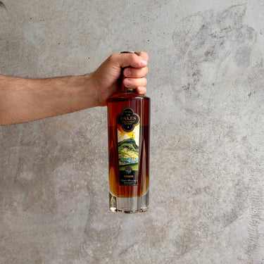 Hand holding a bottle of The Lakes Single Malt Voyage whiskey against a gray concrete wall