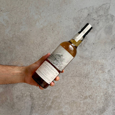 Hand holding a bottle of The Whisky Set against a concrete background