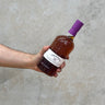 A hand holding a bottle of Tobermory 21 Year Old Oloroso Cask Whisky with a purple cap, against a grey concrete background.