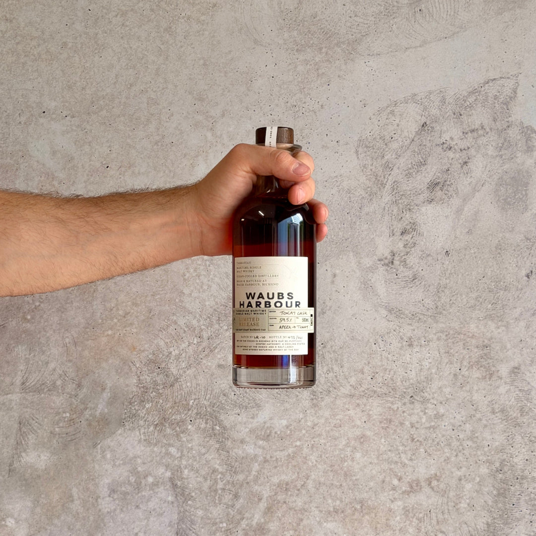 Hand holding a bottle labeled Waubs Harbour Whiskey Tokay Cask against a gray concrete background