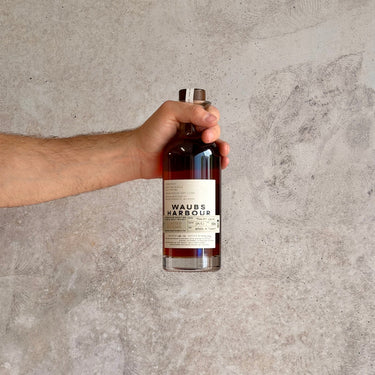 Hand holding a bottle labeled Waubs Harbour Whiskey Tokay Cask against a gray concrete background