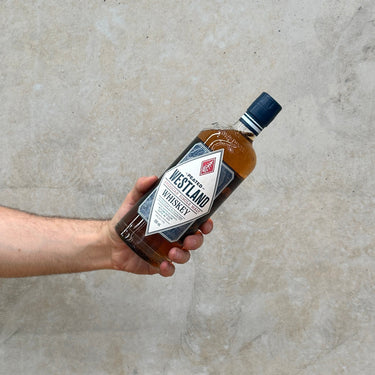 A hand holding a bottle of Westland American Single Malt Peated Whisky with a concrete background.