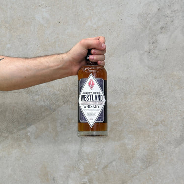 A hand holding a bottle of Westland American Single Malt Sherry Wood whisky against a grey background.