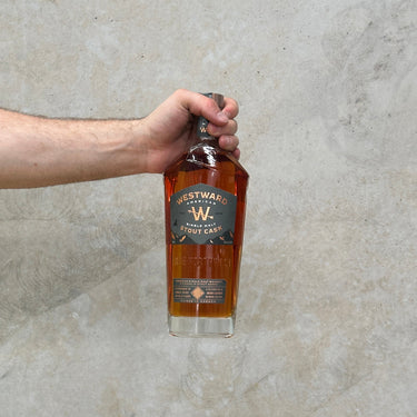 A hand holding a bottle of Westward Stout Cask Whisky against a grey concrete background.