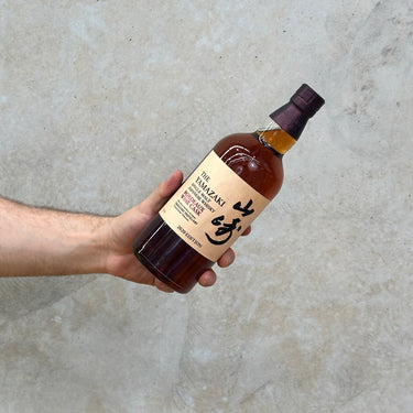 A hand holding a bottle of Yamazaki Bordeaux Wine Cask 2020 Whisky with a label in Japanese script and a clear amber liquid visible.