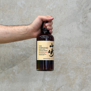 A hand holding a bottle of Yamazaki Spanish Oak 2020 Whisky with a label in Japanese and English on a grey background.