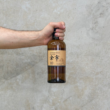 A hand holding a bottle of Yoichi 10 Year Old Single Malt Whisky with a concrete background.