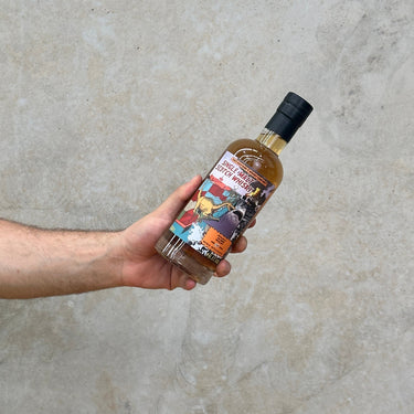 A hand holding a bottle of Aultmore 28 Year Old whisky with a colorful label against a concrete wall.